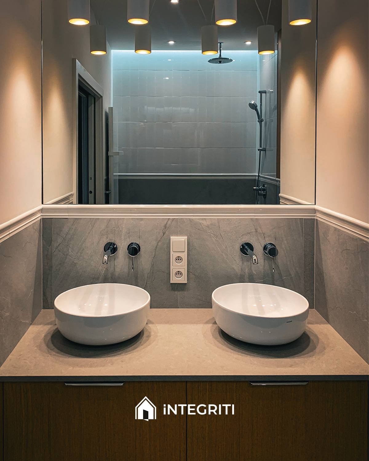 Łazienka z dwoma białymi umywalkami nablatowymi, szarą płytką imitującą kamień na ścianie, bateriami ściennymi w kolorze czarnym, lustrem odbijającym wannę i prysznic oraz oświetleniem punktowym.