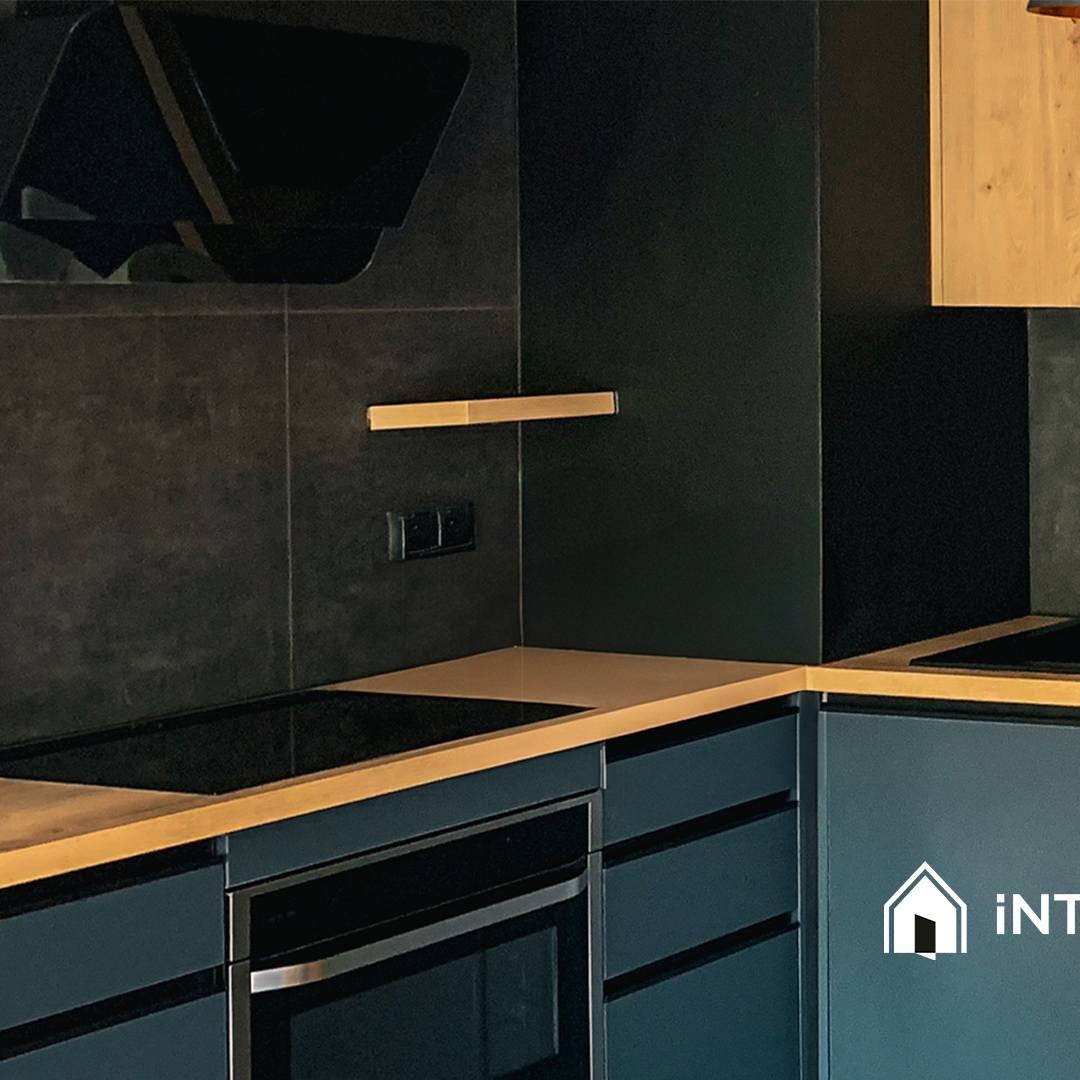 Nowoczesna kuchnia w ciemnych odcieniach z drewnianymi akcentami, płyta indukcyjna i piekarnik w zabudowie, minimalistyczna półka na ścianie, ciemne płytki na ścianie.