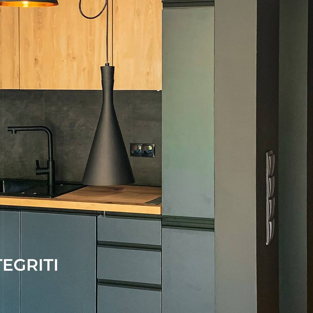 Nowoczesna kuchnia z szarymi szafkami, drewnianym blatem, czarną armaturą i stożkową lampą wiszącą; widoczne gniazdka elektryczne i minimalistyczny uchwyt na ścianie.