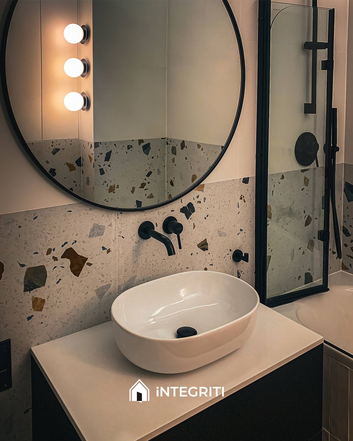 Nowoczesna łazienka z owalną umywalką nablatową, czarną armaturą i lustrem w okrągłej ramie na tle ściany wyłożonej płytkami imitującymi lastryko, z widoczną kabiną prysznicową z czarnymi profilami.