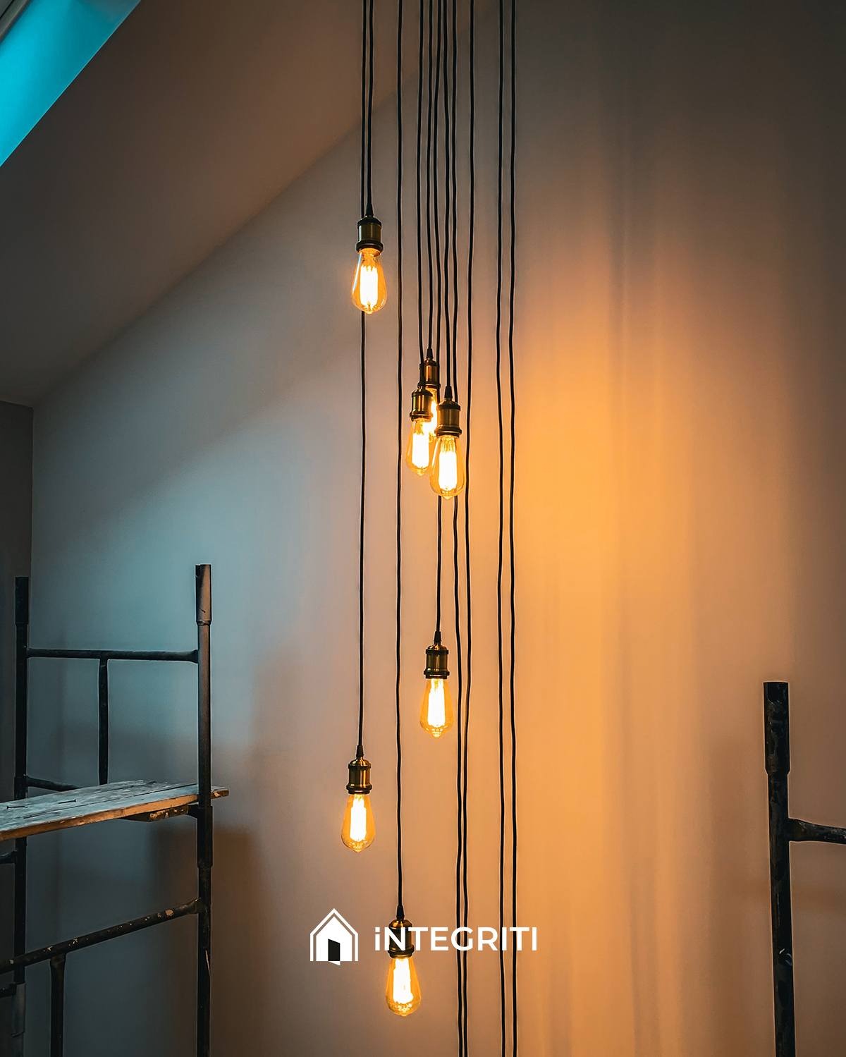 Niedokończone wnętrze z wiszącymi żarówkami Edisona na czarnych kablach różnej długości, widoczne rusztowanie malarskie, logo firmy w rogu.