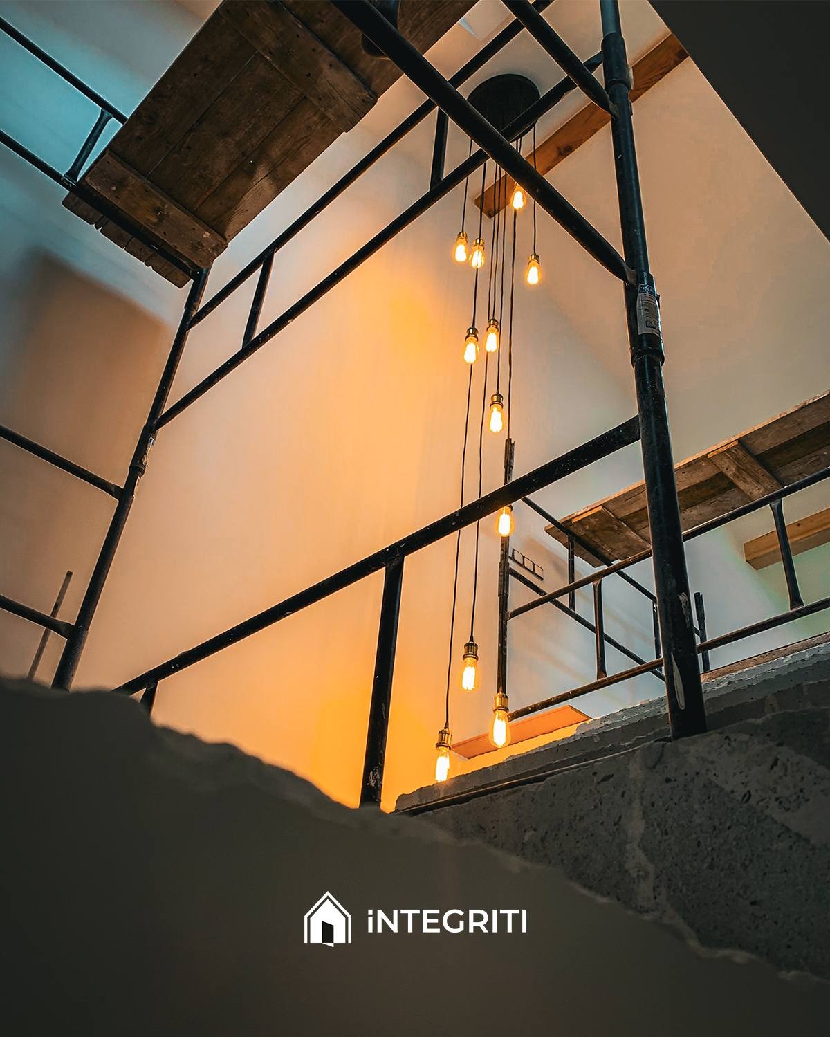 Wnętrze w trakcie prac wykończeniowych z widocznym rusztowaniem, wiszącymi żarówkami dekoracyjnymi i fragmentem surowego betonu, sfotografowane z dołu.