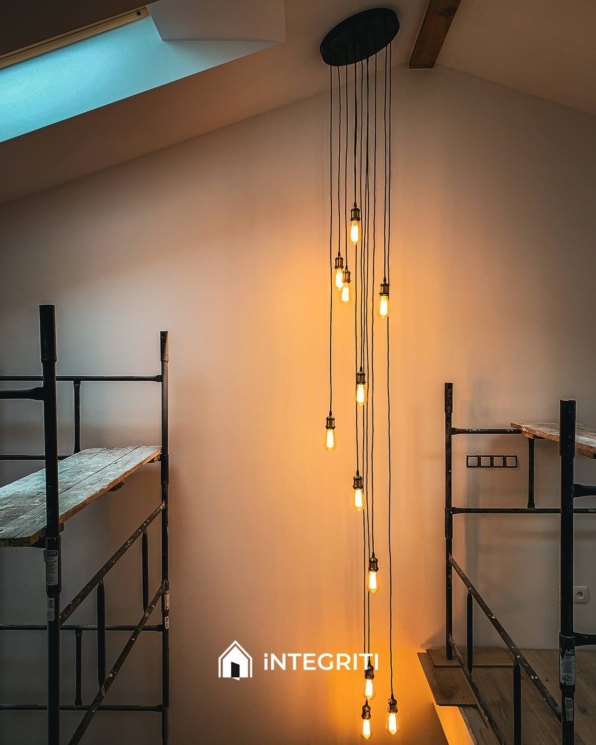 Wysokie pomieszczenie z oknem dachowym, oświetlone wiszącą lampą z żarówkami Edisona, w tle rusztowanie malarskie z drewnianymi podestami, logo firmy w prawym dolnym rogu.