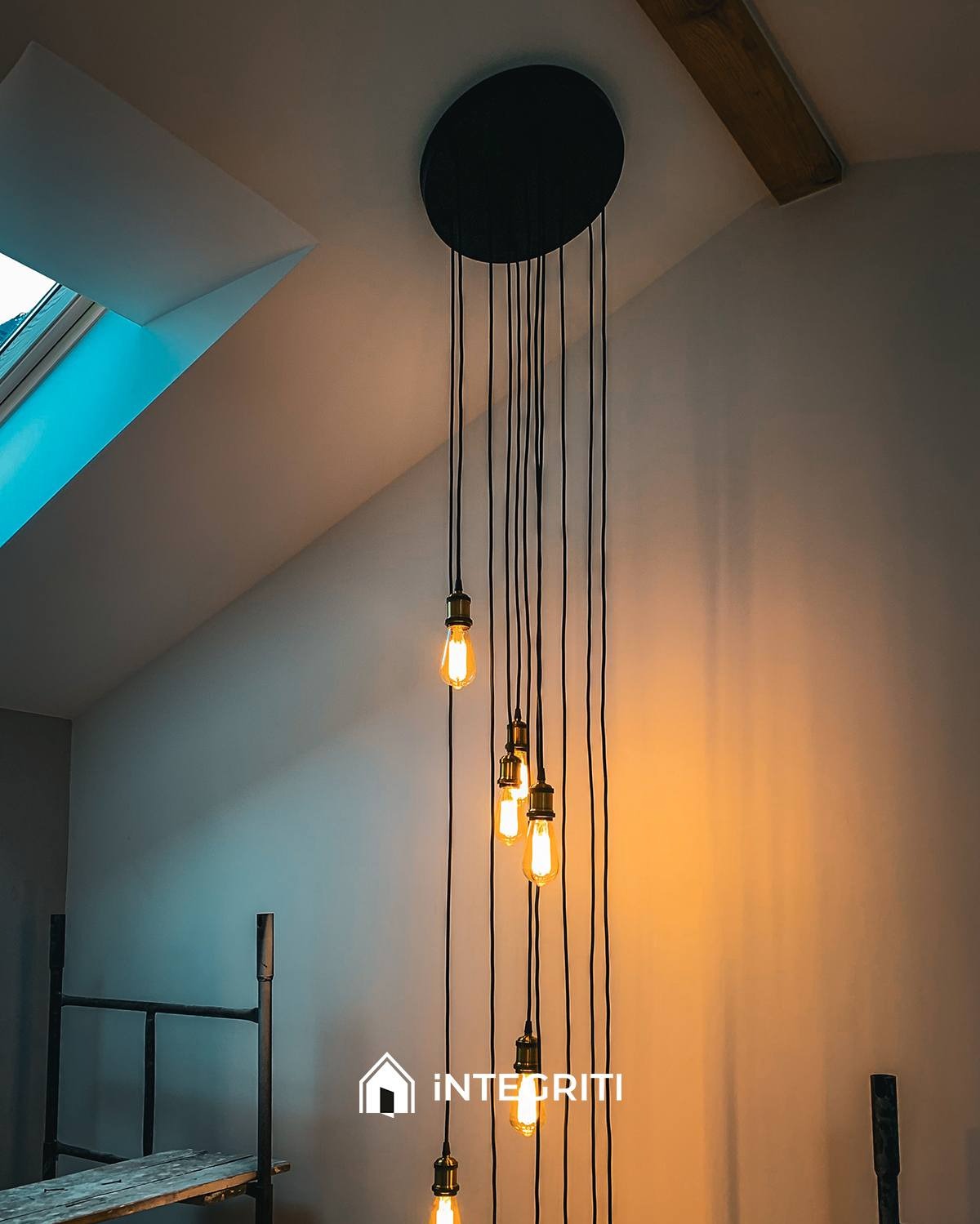 Nierównomiernie rozmieszczone żarówki Edisona zwisające z czarnej, okrągłej podsufitki, widoczne na tle skośnego sufitu poddasza z oknem dachowym i fragmentem rusztowania.