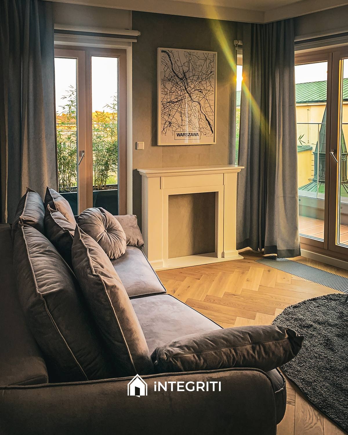 Salon z szarym narożnikiem, parkietem w jodełkę, białym kominkiem i plakatem z mapą Warszawy na ścianie, widok na balkon przez okno.