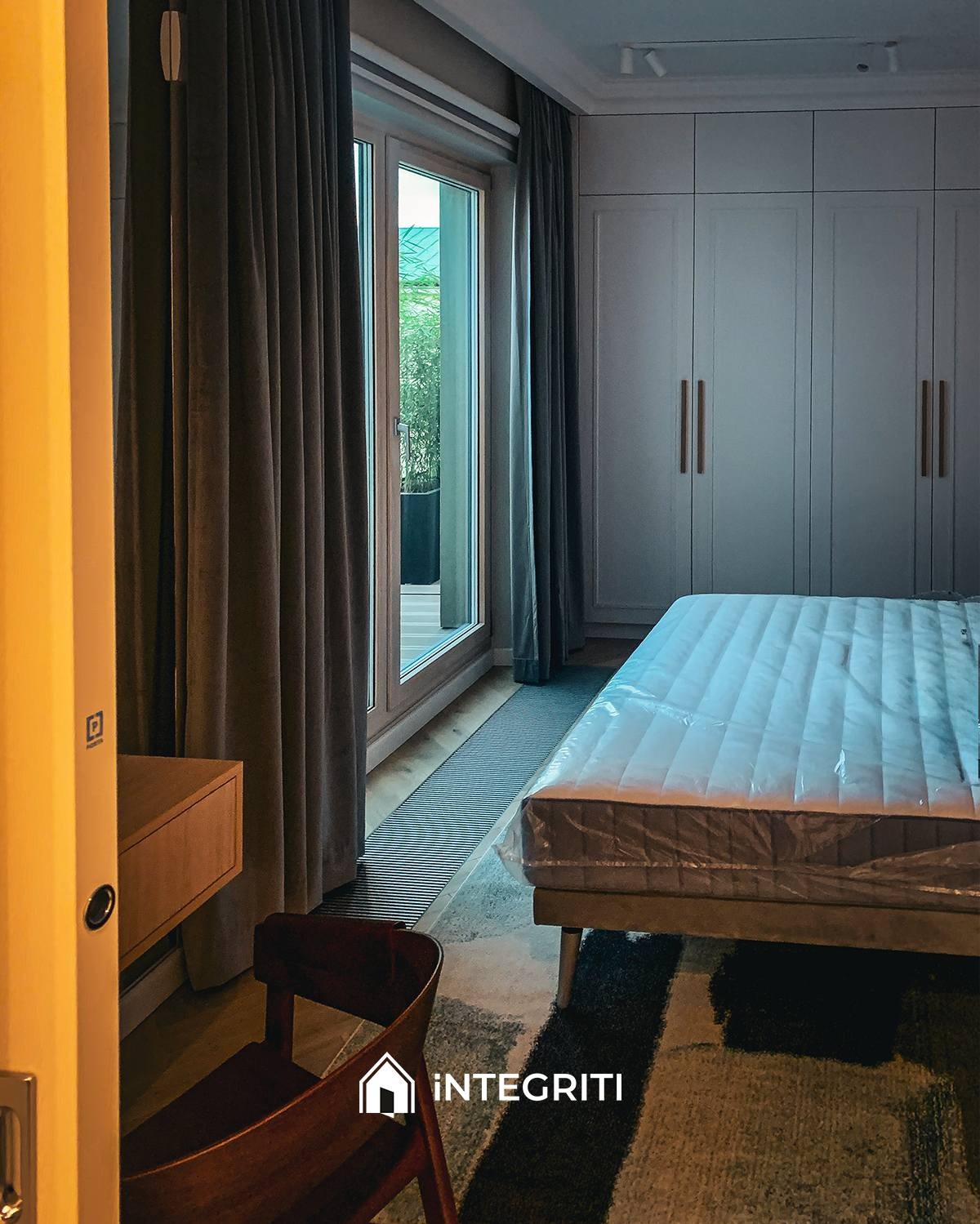 Pokój z łóżkiem w folii ochronnej, szafą wnękową z długimi uchwytami, ciemnymi zasłonami i wyjściem na balkon. Widoczny fragment biurka i krzesła. Na podłodze wzorzysty dywan.