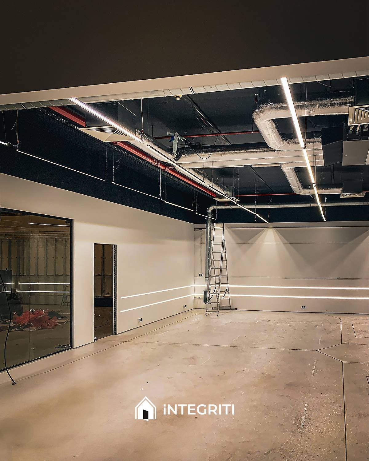 Wnętrze komercyjne w trakcie prac wykończeniowych: surowa podłoga betonowa, białe ściany z poziomymi liniami świetlnymi, czarny sufit z widocznymi instalacjami, drabina oparta o ścianę.