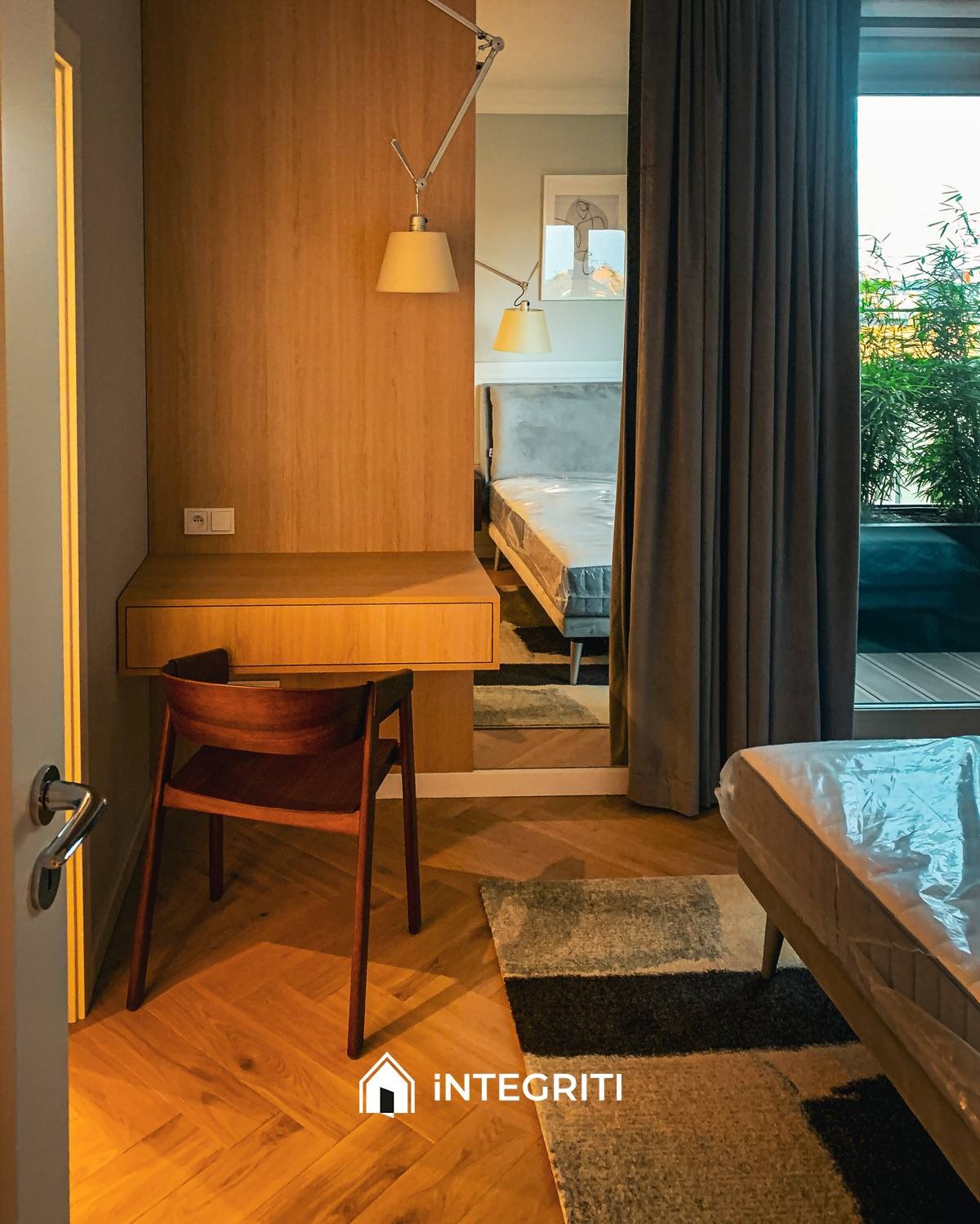 Nowoczesny pokój z drewnianą podłogą w jodełkę, biurkiem z wbudowaną szafką, krzesłem i łóżkiem zabezpieczonym folią, widok przez otwarte drzwi.