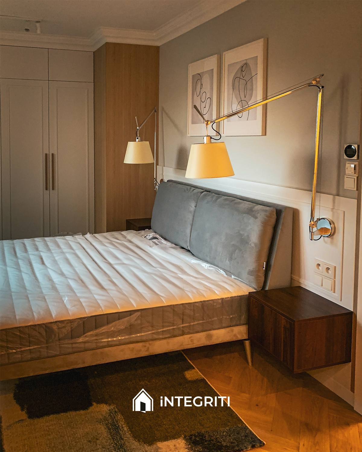 Nowoczesna sypialnia z szarą tapicerowaną ramą łóżka, materacem w folii, drewnianymi szafkami nocnymi, regulowanymi lampami ściennymi i abstrakcyjnymi obrazami na ścianie.