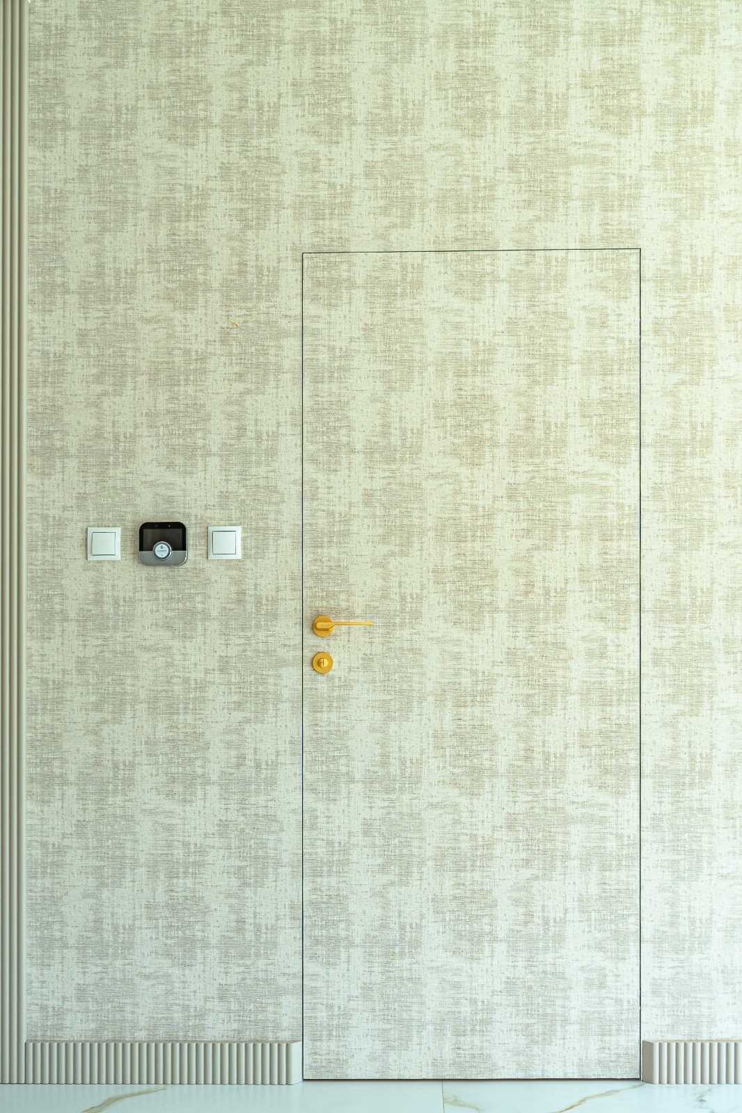 Beżowa ściana z ukrytymi drzwiami i złotą klamką, obok włączniki światła i panel sterowania. Minimalistyczny design wnętrza z subtelną fakturą tapety.