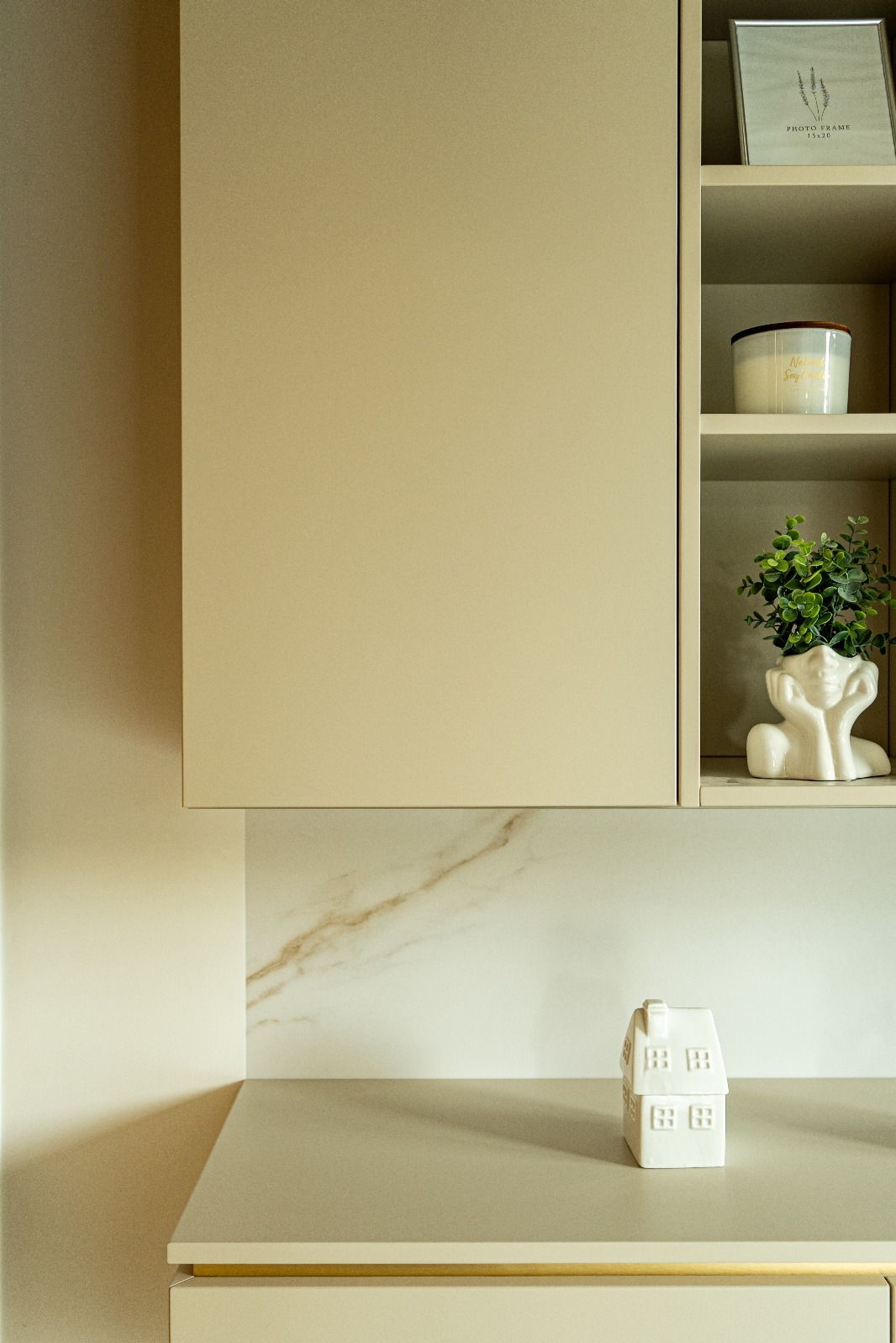 Beżowa zabudowa kuchenna z marmurowym blatem i dekoracjami: ramka, świeca, doniczka w kształcie głowy i ceramiczny domek. Minimalistyczny design.
