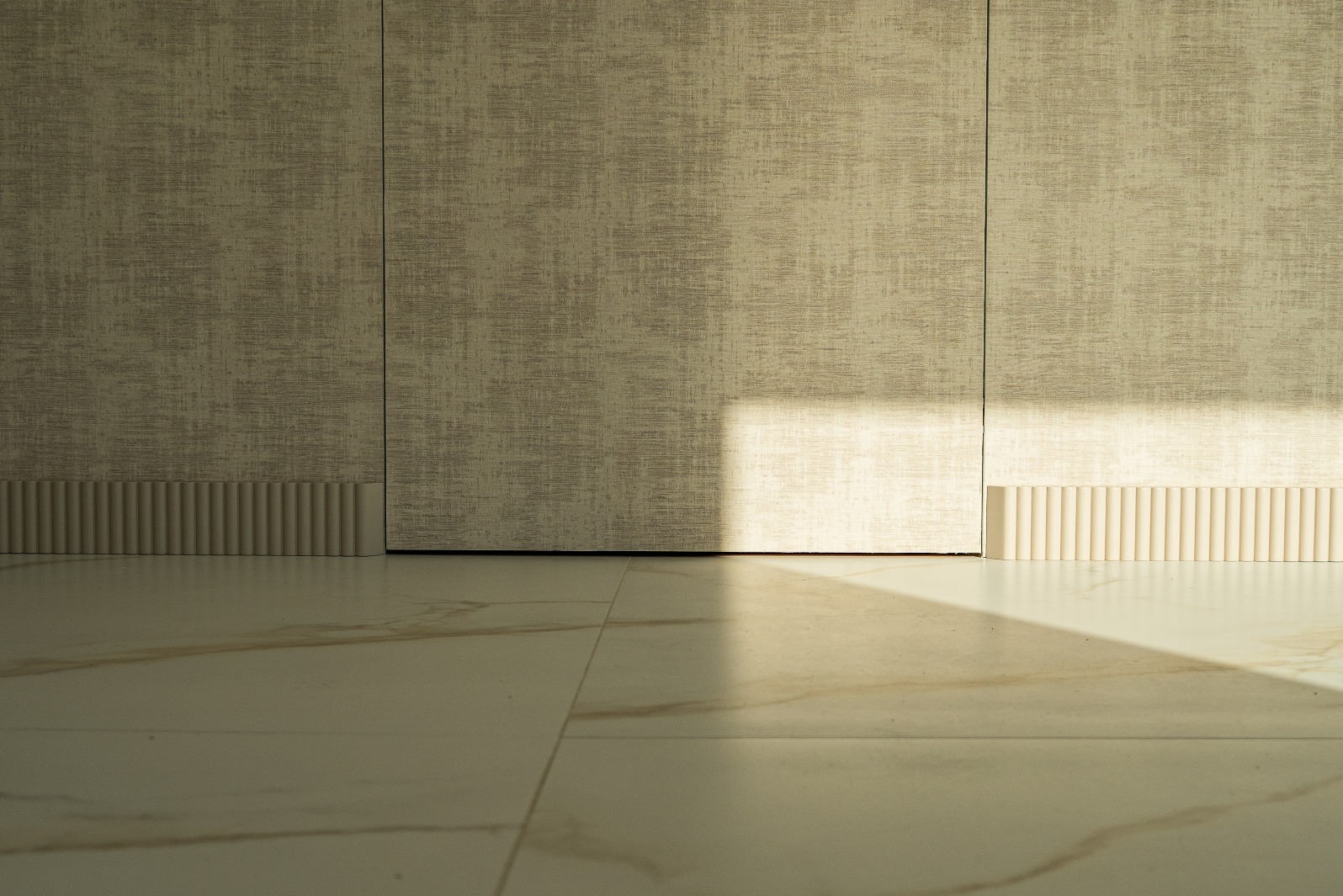 Beżowa ściana z dekoracyjną listwą przypodłogową i marmurową podłogą, oświetlona naturalnym światłem tworzącym kontrastowy cień. Minimalistyczne wykończenie wnętrza.