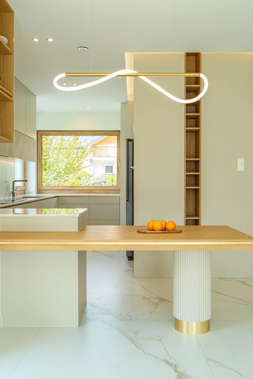 Nowoczesna kuchnia z wyspą i oświetleniem LED. Na blacie pomarańcze, w tle okno z widokiem na zieleń. Marmurowe płytki podłogowe i minimalistyczne szafki.