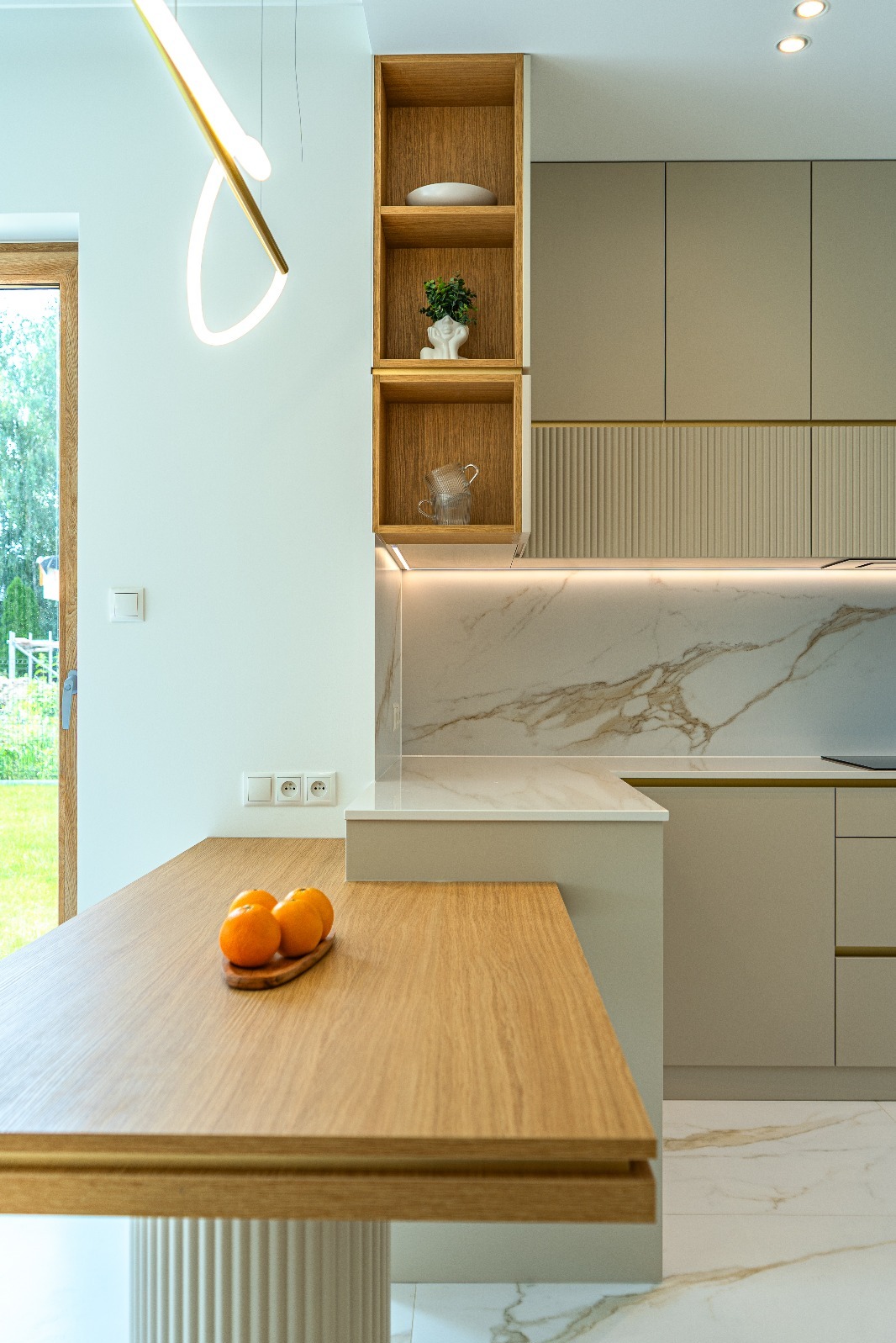 Nowoczesna kuchnia z drewnianym blatem, marmurowym backsplash'em i otwartymi półkami z dekoracjami. Widok na ogród przez okno. Oświetlenie LED podkreśla detale.