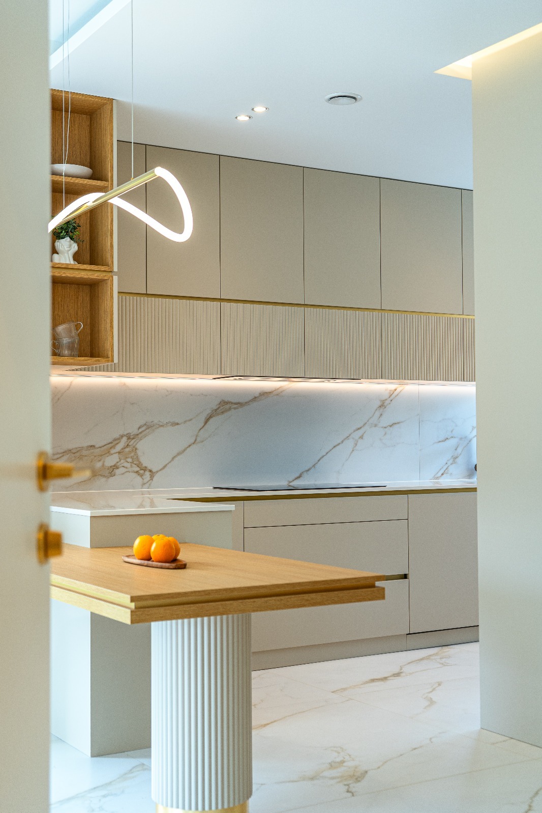 Elegancka kuchnia w beżach z marmurowym blatem i złotymi akcentami. Na drewnianym stole leżą pomarańcze. Widok zza uchylonych drzwi.