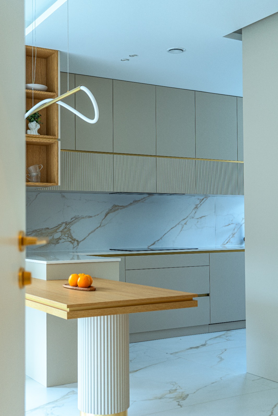 Nowoczesna kuchnia w odcieniach szarości z marmurowym blatem i drewnianym stołem z owocami, widziana zza uchylonych drzwi. Minimalistyczny design i subtelne oświetlenie.