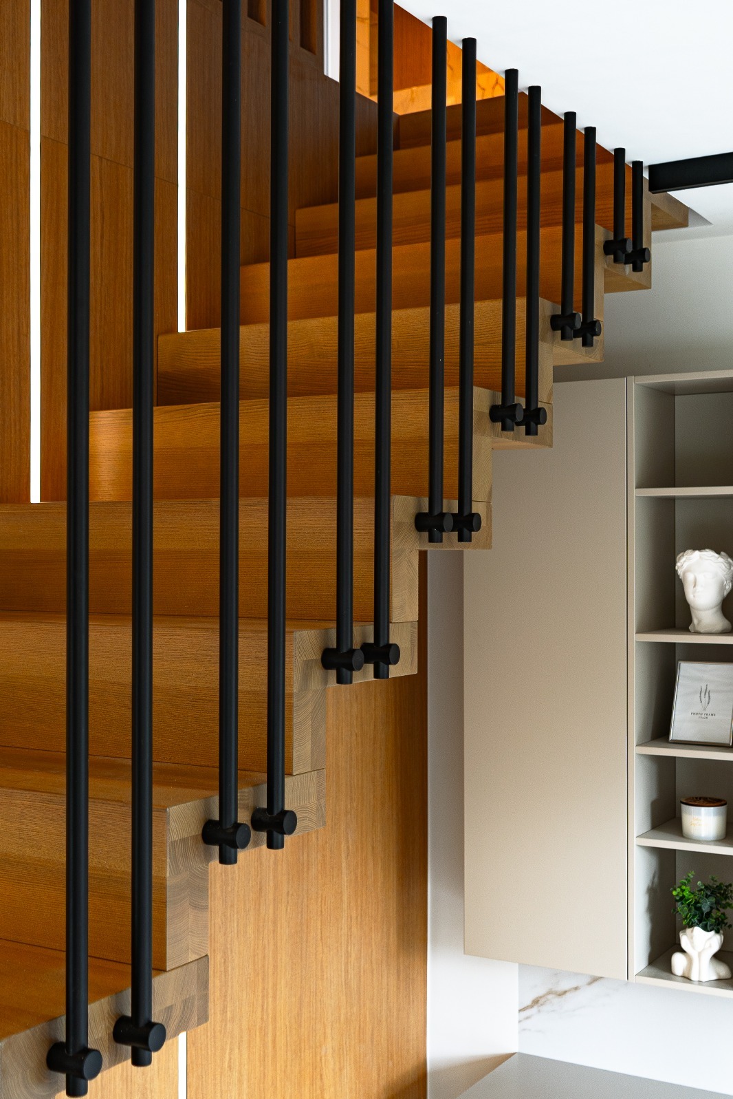 Drewniane schody z czarną, minimalistyczną balustradą i subtelnym oświetleniem LED, uzupełnione szafką z dekoracjami w tle. Nowoczesny design.