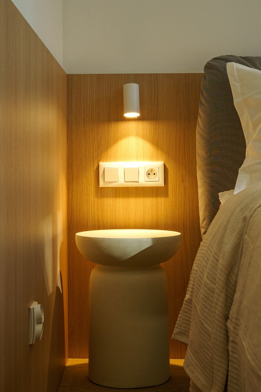 Drewniana ściana z włącznikami i gniazdkiem oświetlona lampką. Obok stolik nocny i fragment łóżka. Ciepłe oświetlenie wnętrza.