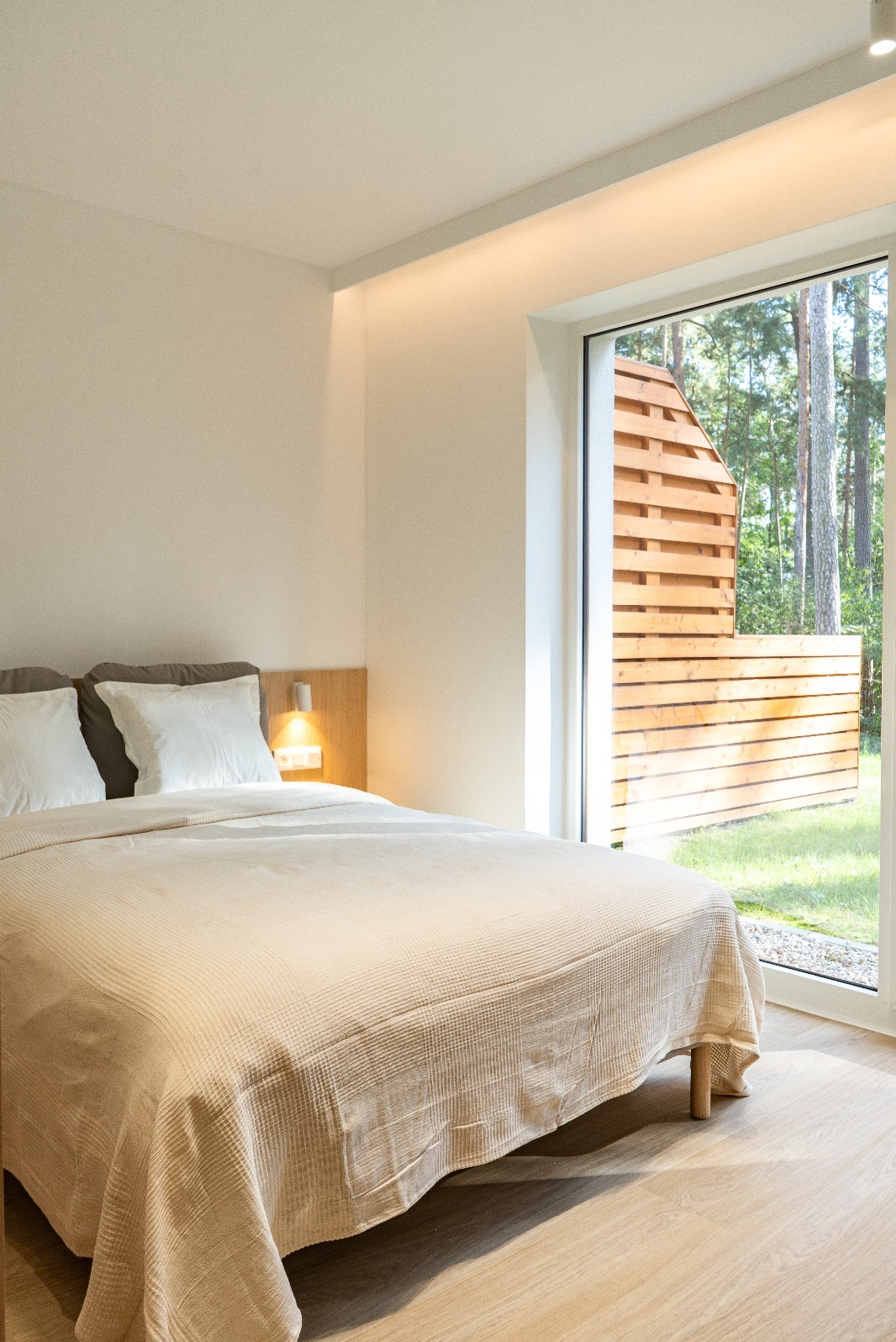 Jasna sypialnia z dużym oknem na ogród. Widoczne łóżko z beżową narzutą, drewniana podłoga i oświetlenie punktowe. Minimalistyczny design wnętrza.