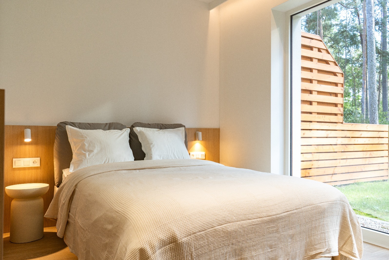 Jasna sypialnia z dużym oknem na zewnątrz. Widok na zielony ogród i drewniany płot. Minimalistyczny wystrój, beżowa narzuta na łóżku, lampki nocne.