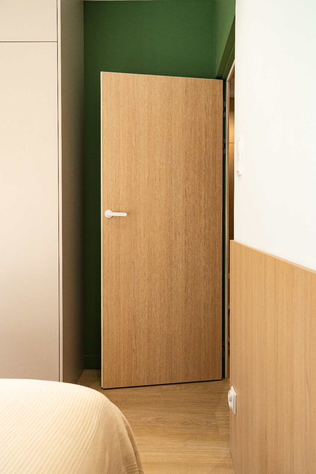 Otwarta, fornirowana dębem drzwi wewnętrzne z białą klamką, na tle zielonej ściany i beżowej szafy w minimalistycznym wnętrzu. Widoczny fragment łóżka.