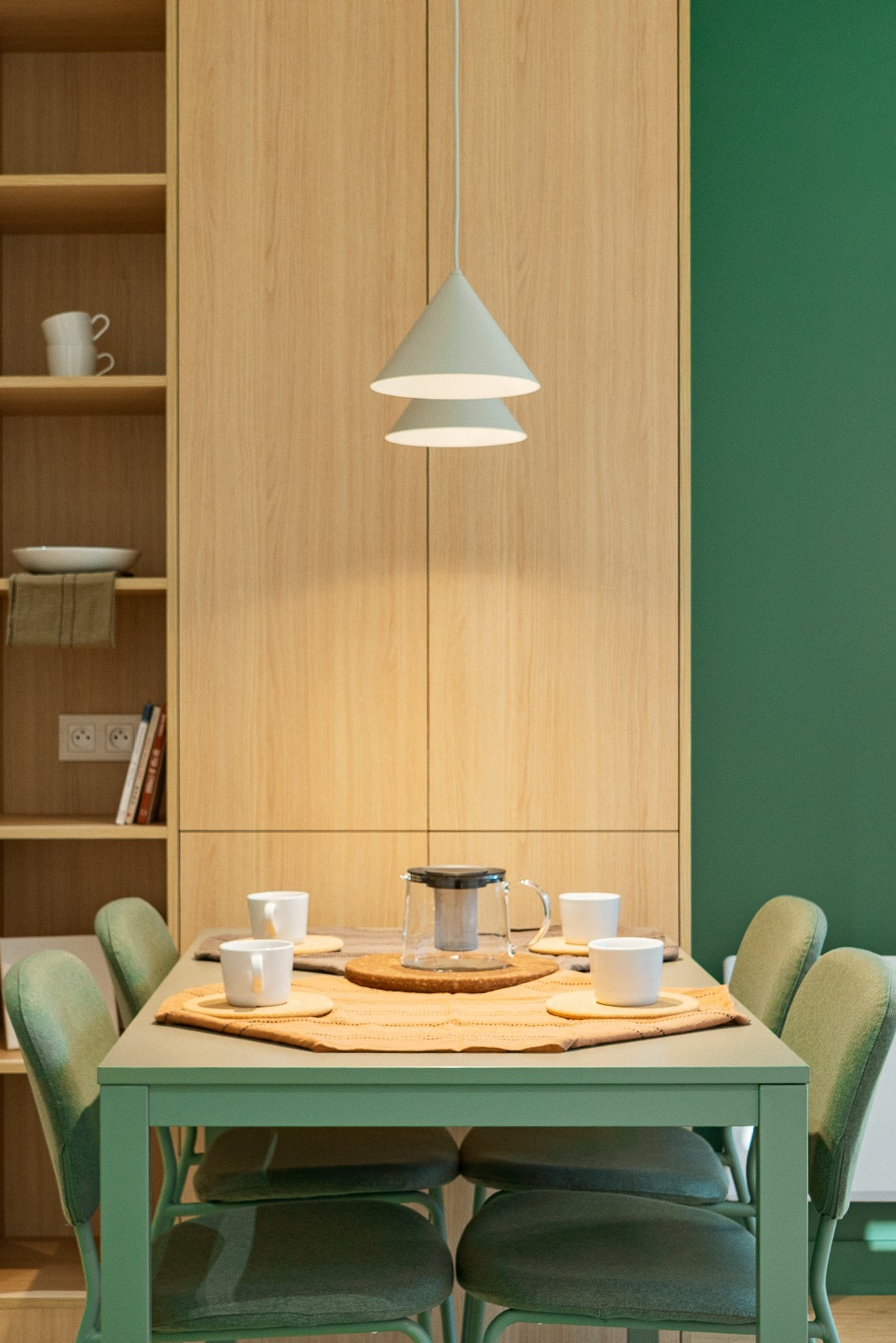 Stylowa jadalnia z zielonymi akcentami: ściana, krzesła i stół w odcieniach zieleni. Drewniane szafki w tle i nowoczesna lampa nad stołem. Minimalistyczny design.