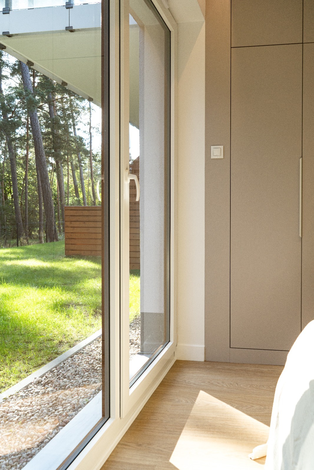 Nowoczesne okno z widokiem na zieleń, w minimalistycznym wnętrzu z drewnianą podłogą i zabudowaną szafą w odcieniach beżu. Jasne, naturalne światło.