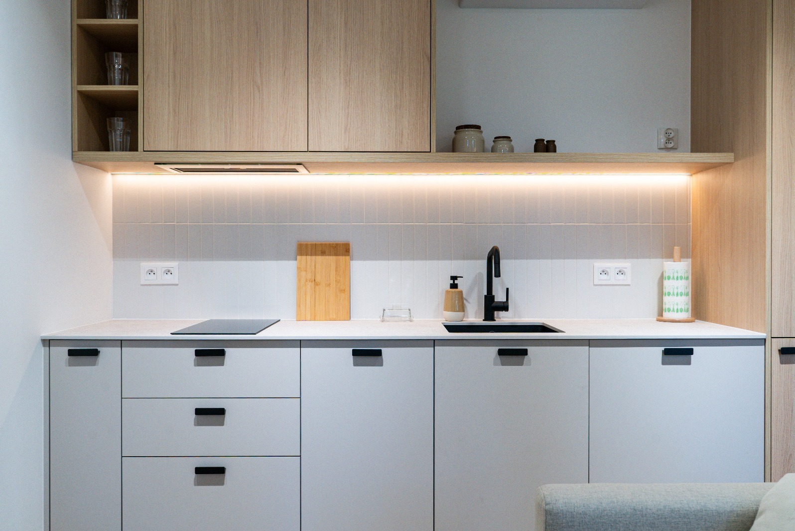 Nowoczesna kuchnia z białymi szafkami, drewnianymi akcentami i czarną armaturą. Minimalistyczny design z oświetleniem LED pod szafkami wiszącymi.