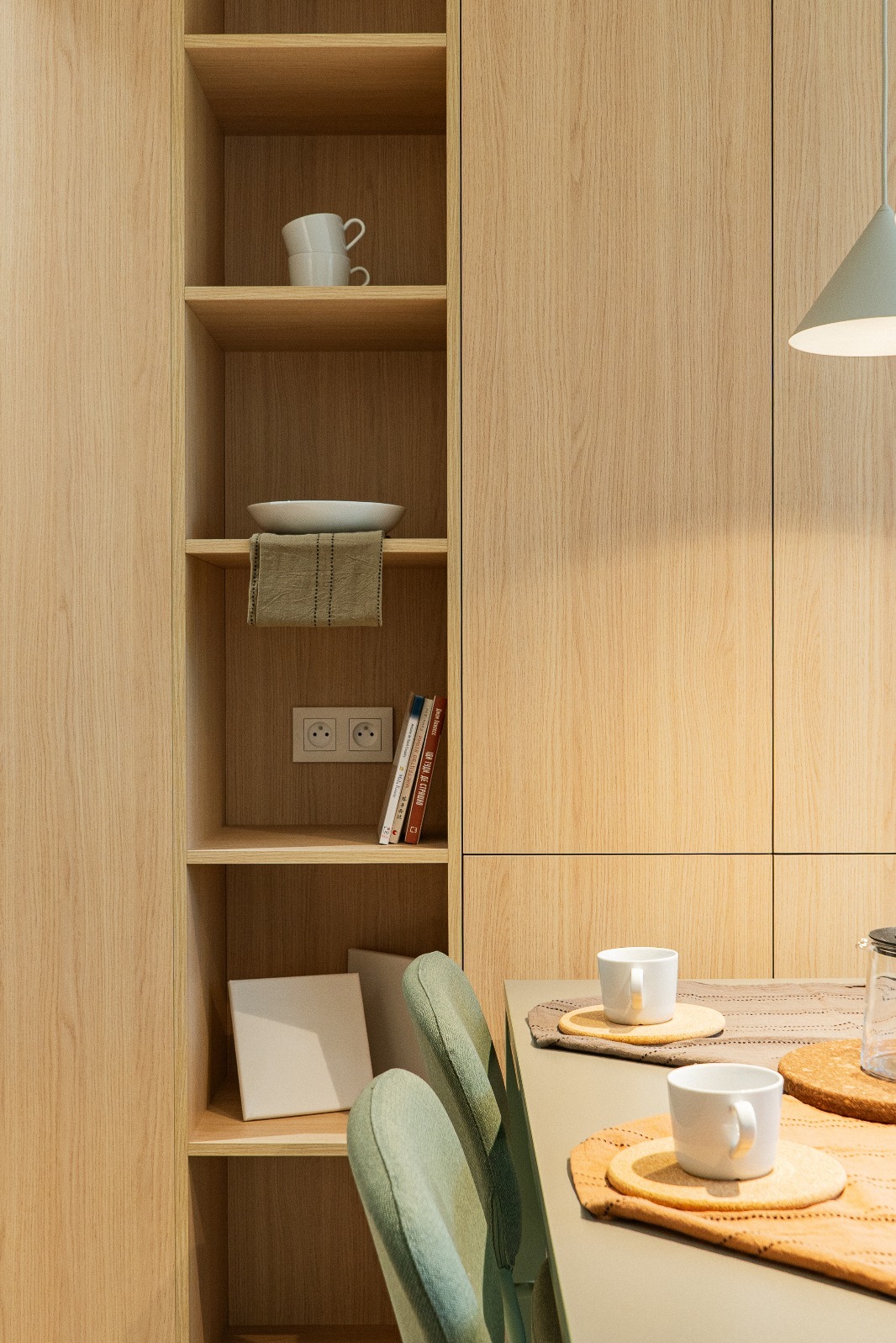 Jasne, drewniane szafki kuchenne z otwartymi półkami, na których stoją naczynia i książki. Na pierwszym planie stół z filiżankami i krzesła.