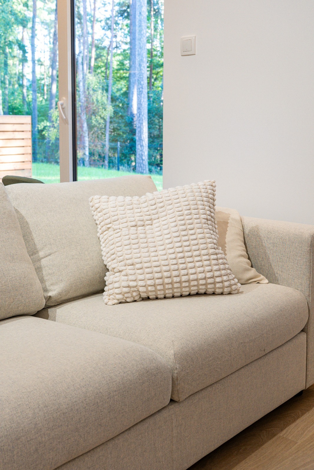 Jasna sofa z dekoracyjną poduszką w minimalistycznym wnętrzu z widokiem na las przez okno. Neutralna kolorystyka, nowoczesny design.
