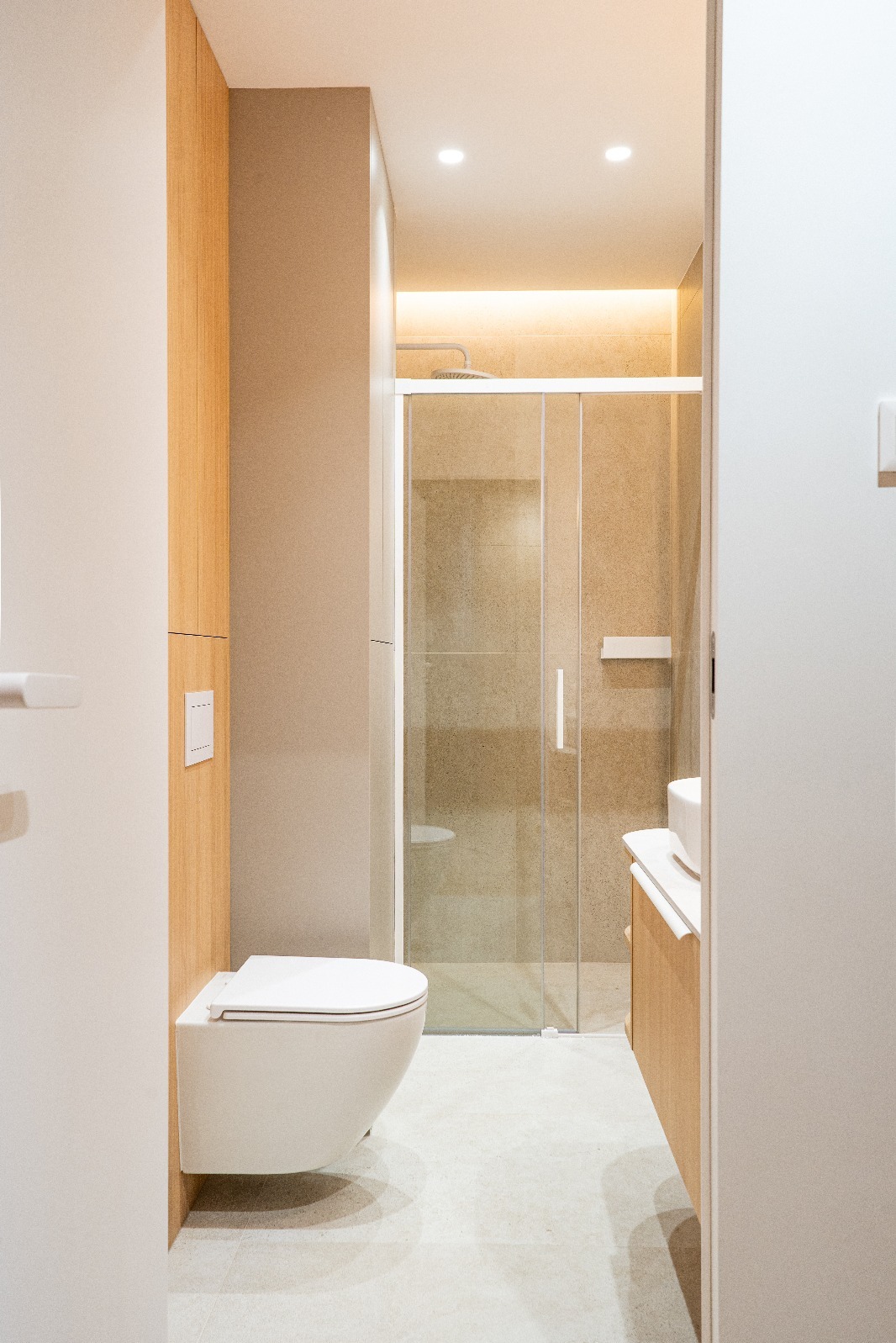 Nowoczesna łazienka z podwieszaną toaletą, kabiną prysznicową ze szklanymi drzwiami i drewnianymi akcentami, oświetlona ciepłym światłem.