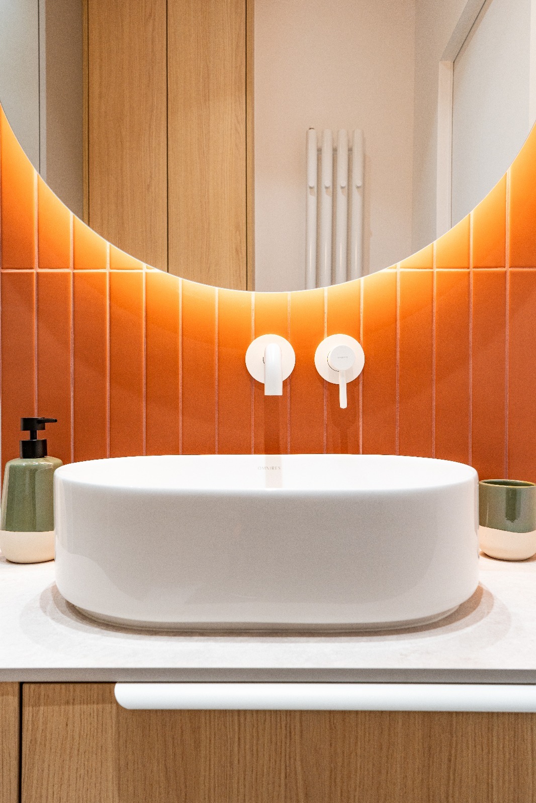 Nowoczesna łazienka z owalną umywalką nablatową, pomarańczowymi płytkami i podświetlanym lustrem. Minimalistyczne baterie i akcesoria w odcieniach bieli i zieleni.