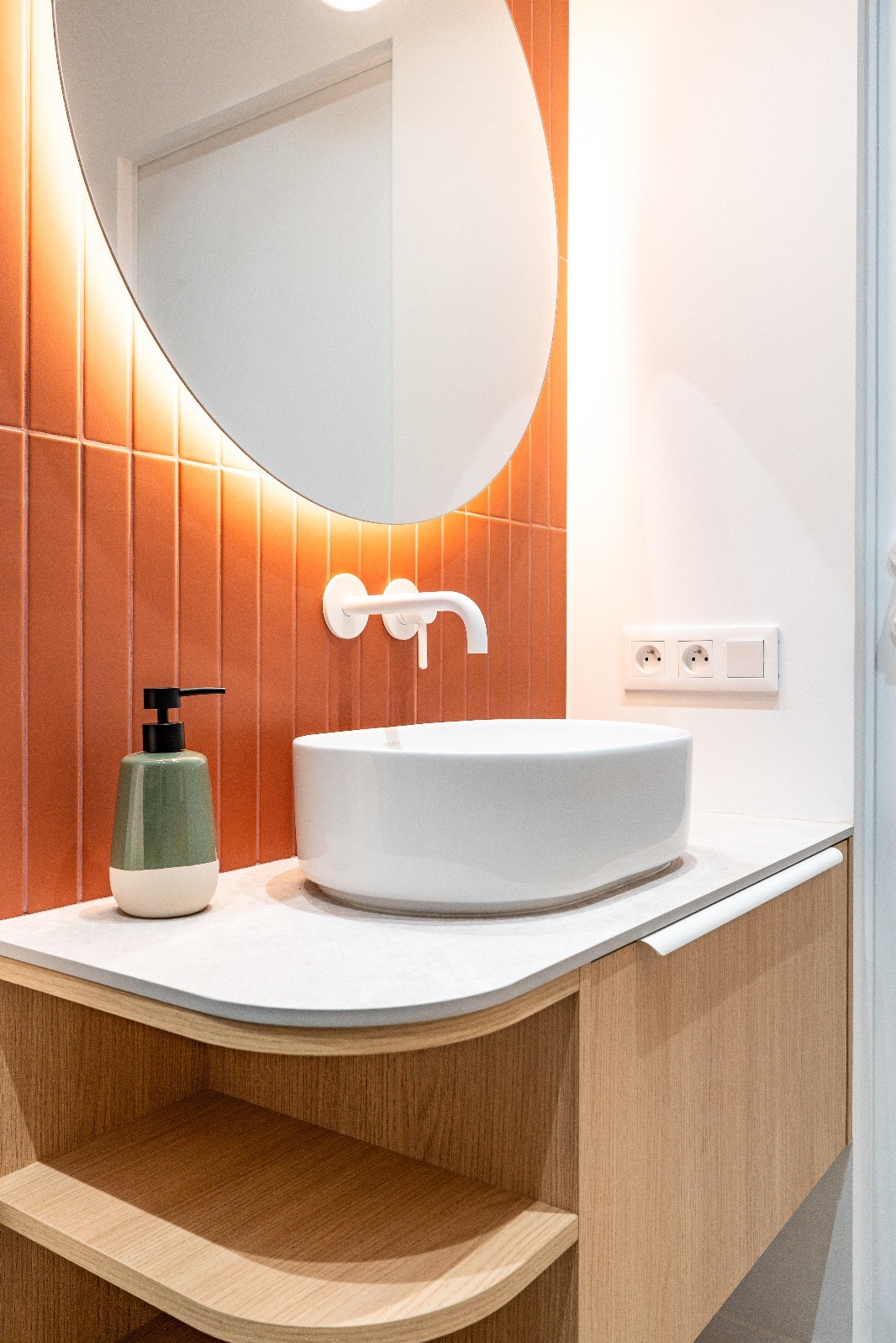 Nowoczesna łazienka z okrągłym lustrem podświetlonym LED, umywalką nablatową i pomarańczowymi płytkami ułożonymi pionowo. Minimalistyczny design, ciepłe oświetlenie.