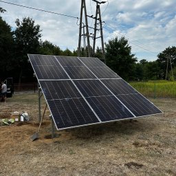 Nex Energy - Montaż paneli fotowoltaicznych na konstrukcji wsporczej na trawiastym terenie, widoczna łopata i materiały montażowe, słup energetyczny w tle.