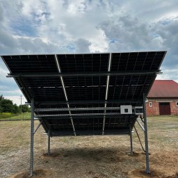 Nex Energy - Widok od spodu na zainstalowane panele słoneczne na metalowej konstrukcji, z widocznym inwerterem i okablowaniem, na tle budynku gospodarczego z czerwonym dachem i pochmurnego nieba.