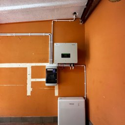 Nex Energy - Montaż instalacji z inwerterem i skrzynką zabezpieczeń na pomarańczowej ścianie w pomieszczeniu z brązową podłogą. Widoczny także akumulator energii.