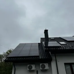 Panele słoneczne na dachu domu z czarną dachówką, widoczne dwa klimatyzatory Kaisai na białej elewacji pod panelami, pochmurne niebo.