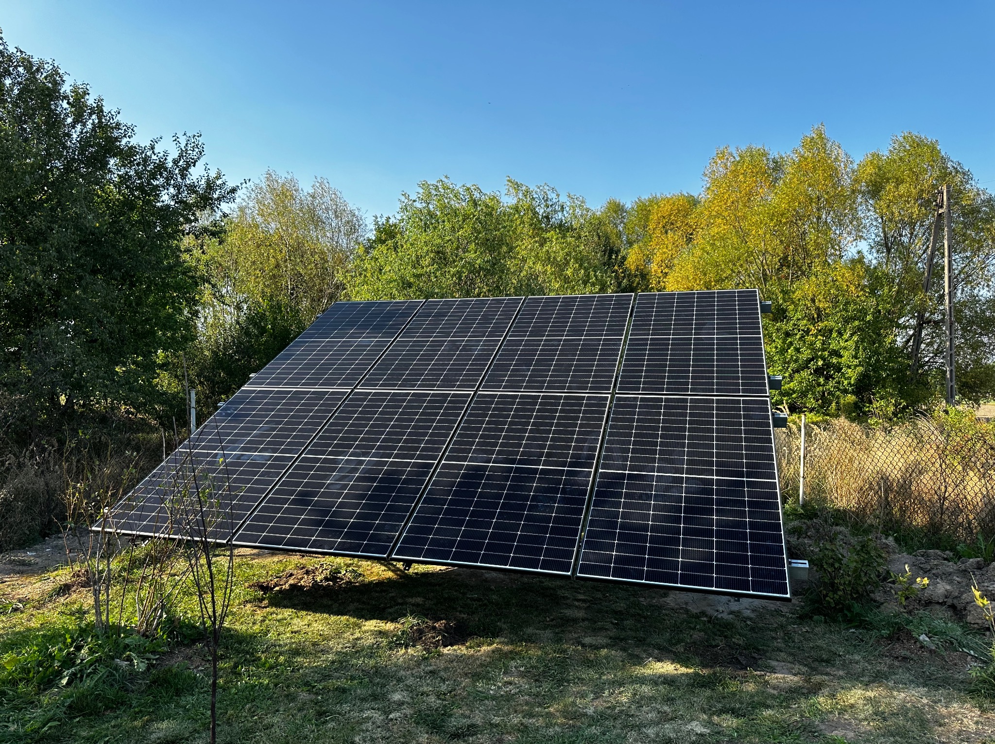 Instalacja paneli słonecznych na stelażu w ogrodzie, widoczne drzewa w tle pod błękitnym niebem.