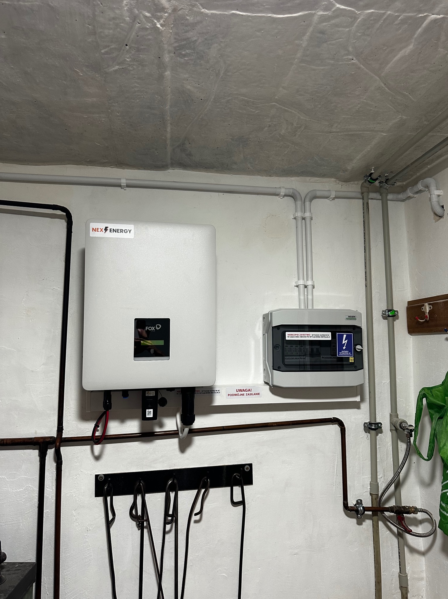 Zamontowany inwerter marki FOX oraz rozdzielnia PV-AC w pomieszczeniu technicznym z instalacjami i rurami na ścianach. Na inwerterze naklejka z logo NEX ENERGY.