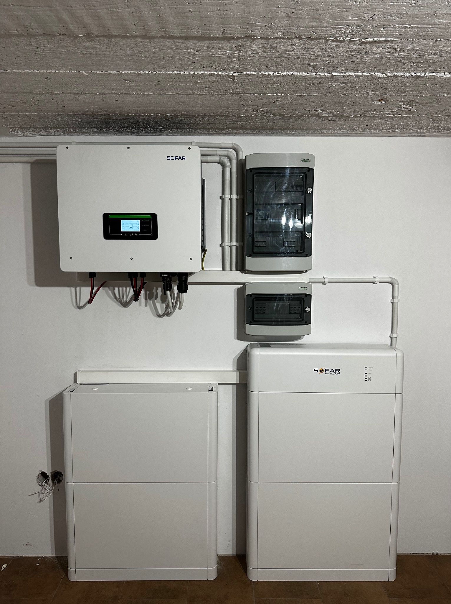 Kompletna instalacja zasilania awaryjnego z inwerterem i dwoma modułami bateryjnymi SOFAR, zabezpieczeniami oraz okablowaniem w piwnicy z surowym betonowym sufitem.