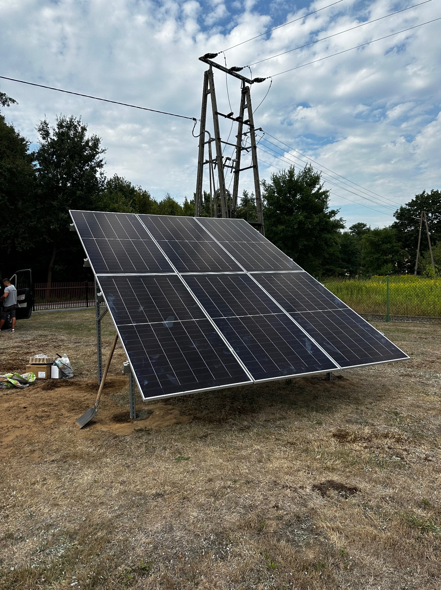 Montaż paneli fotowoltaicznych na konstrukcji wsporczej na trawiastym terenie, widoczna łopata i materiały montażowe, słup energetyczny w tle.