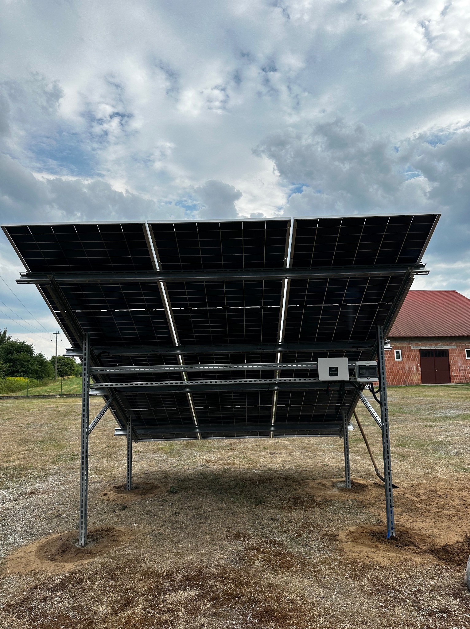 Widok od spodu na zainstalowane panele słoneczne na metalowej konstrukcji, z widocznym inwerterem i okablowaniem, na tle budynku gospodarczego z czerwonym dachem i pochmurnego nieba.