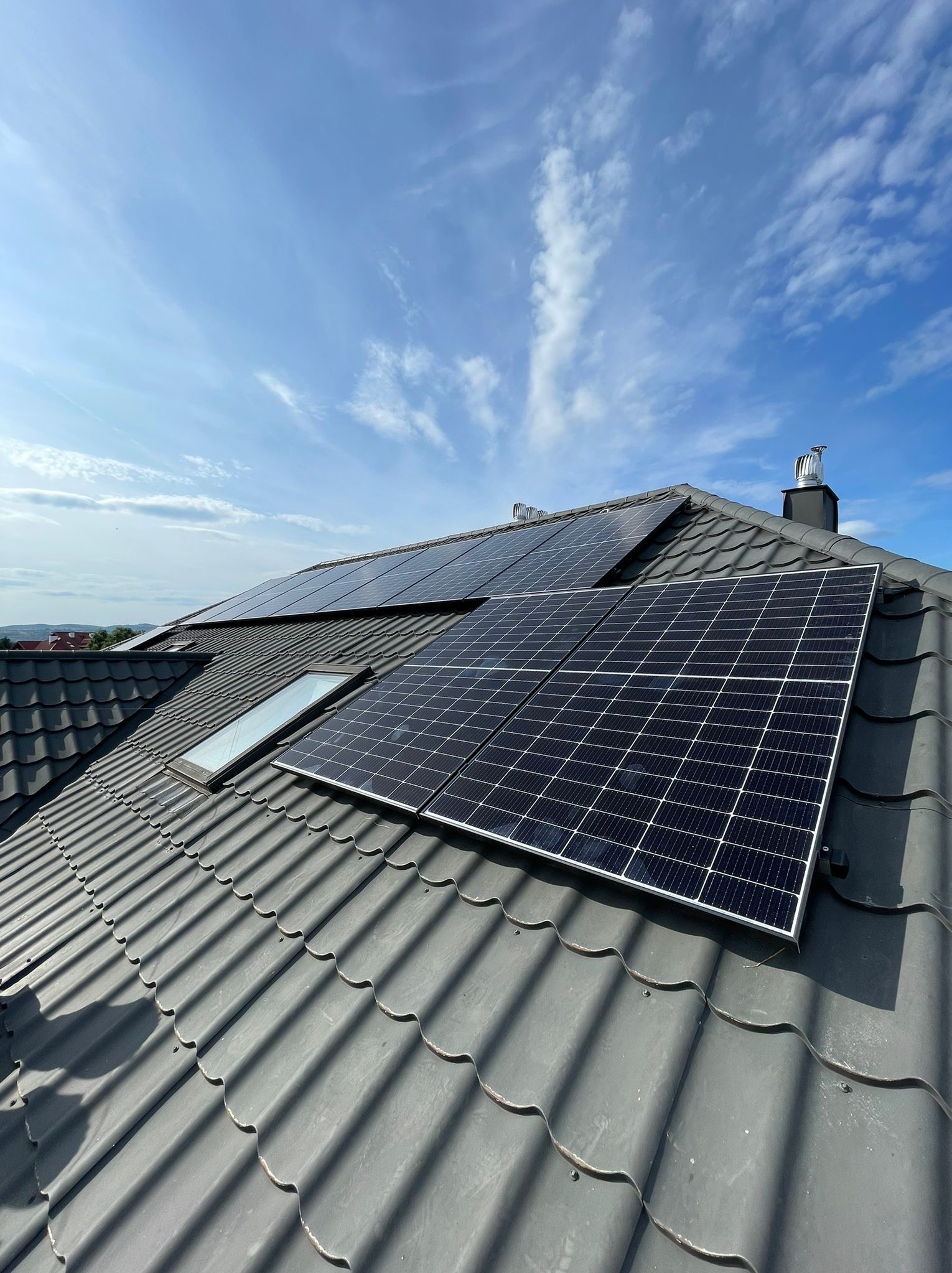 Panele na dachu z dachówką w odcieniach szarości, widoczne okno dachowe i komin, błękitne niebo z chmurami w tle.