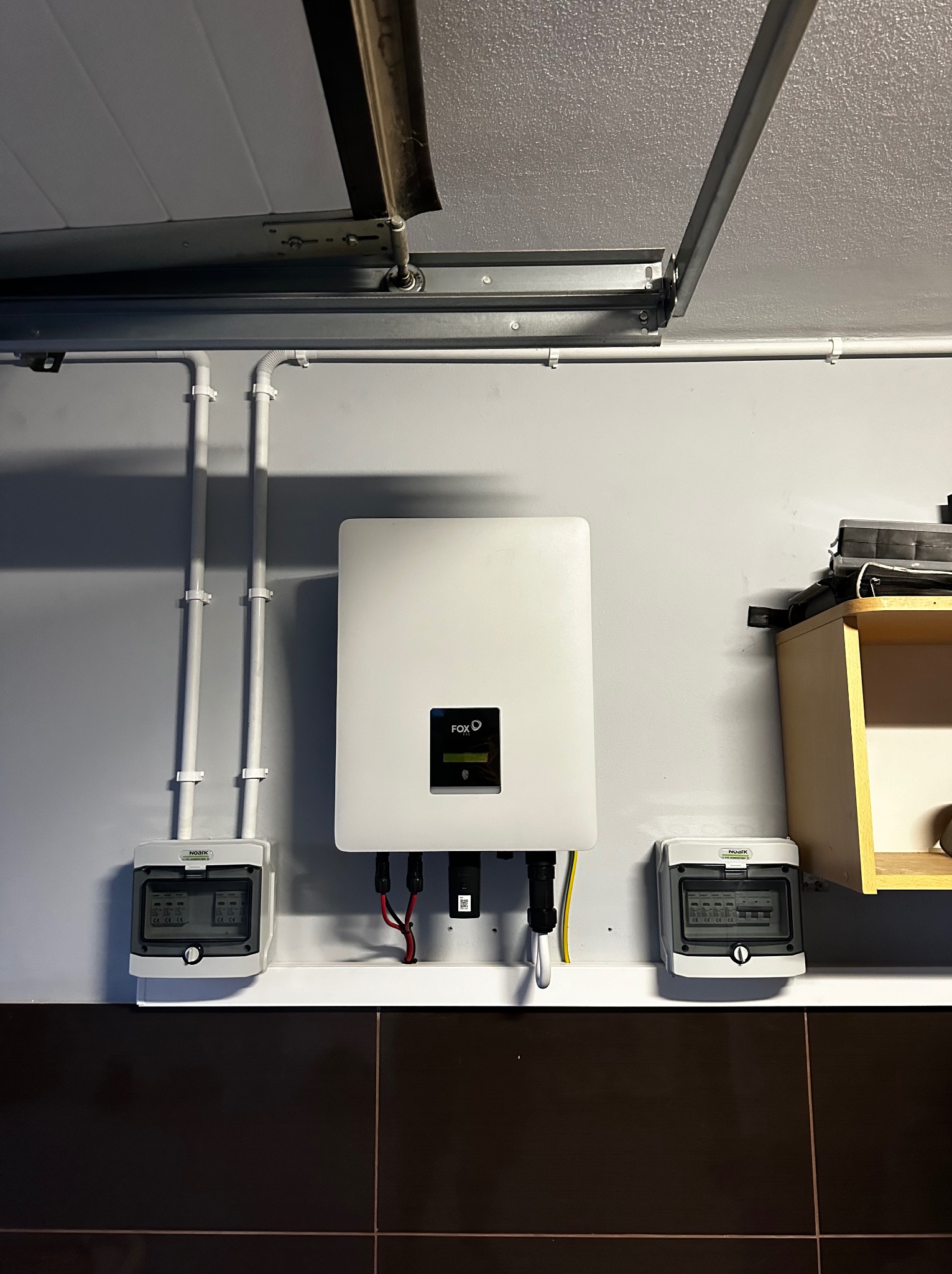 Biała obudowa falownika FOX ESS zamontowana na szarej ścianie w garażu, z widocznymi przewodami, zabezpieczeniami i instalacją elektryczną.