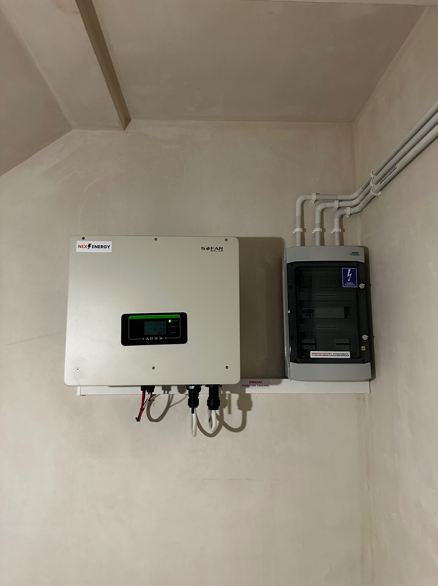 Zamontowany inwerter z logotypem Nex Energy i Sofar oraz skrzynka rozdzielcza PV-DC i PV-AC na ścianie wewnątrz budynku, widoczne przewody i rury instalacyjne.