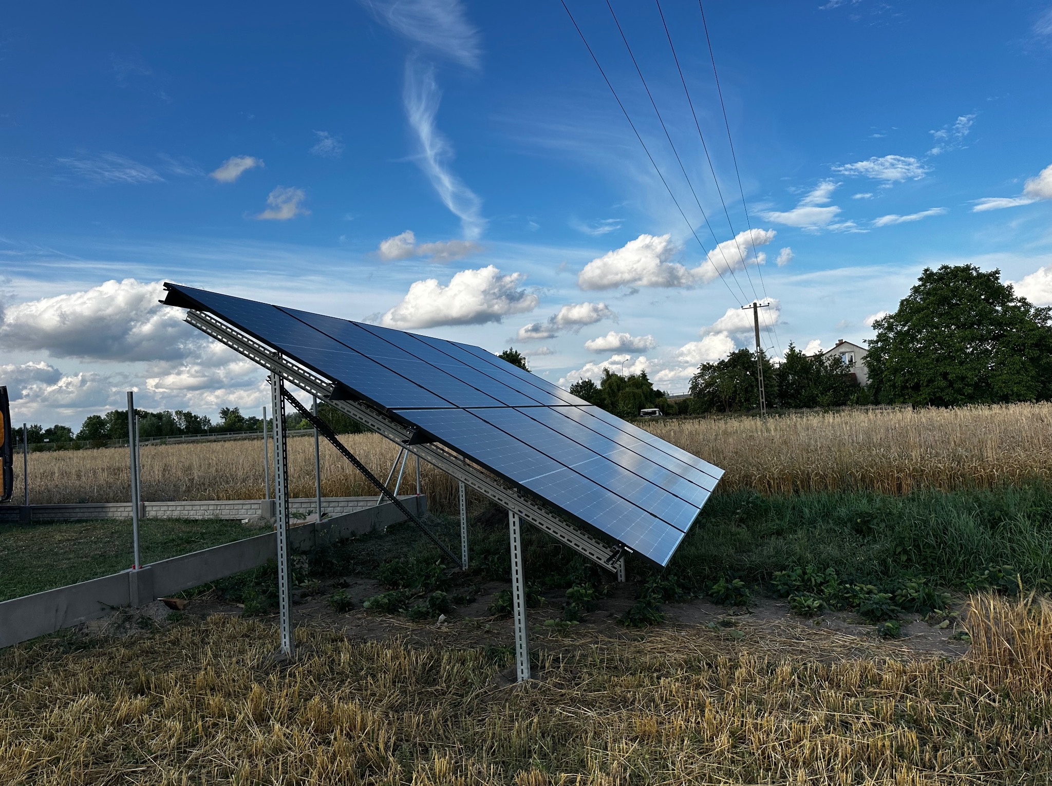 Panel fotowoltaiczny ustawiony na metalowej konstrukcji na polu ze ściętym zbożem, z widoczną linią energetyczną i błękitnym niebem z chmurami w tle.