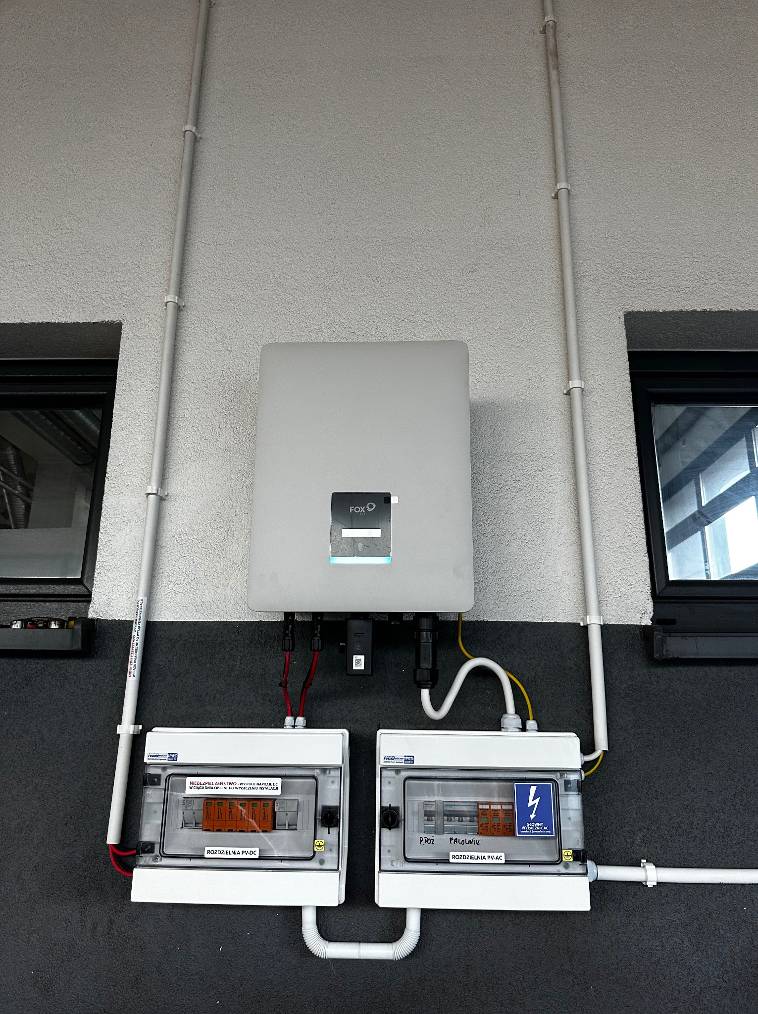 Szary inwerter FoxESS i dwie białe rozdzielnice PV-DC i PV-AC zamontowane na szarej elewacji budynku, z widocznymi przewodami i rurkami instalacyjnymi.