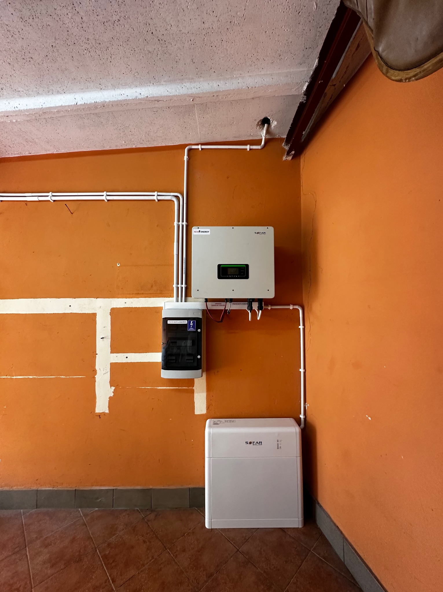 Montaż instalacji z inwerterem i skrzynką zabezpieczeń na pomarańczowej ścianie w pomieszczeniu z brązową podłogą. Widoczny także akumulator energii.