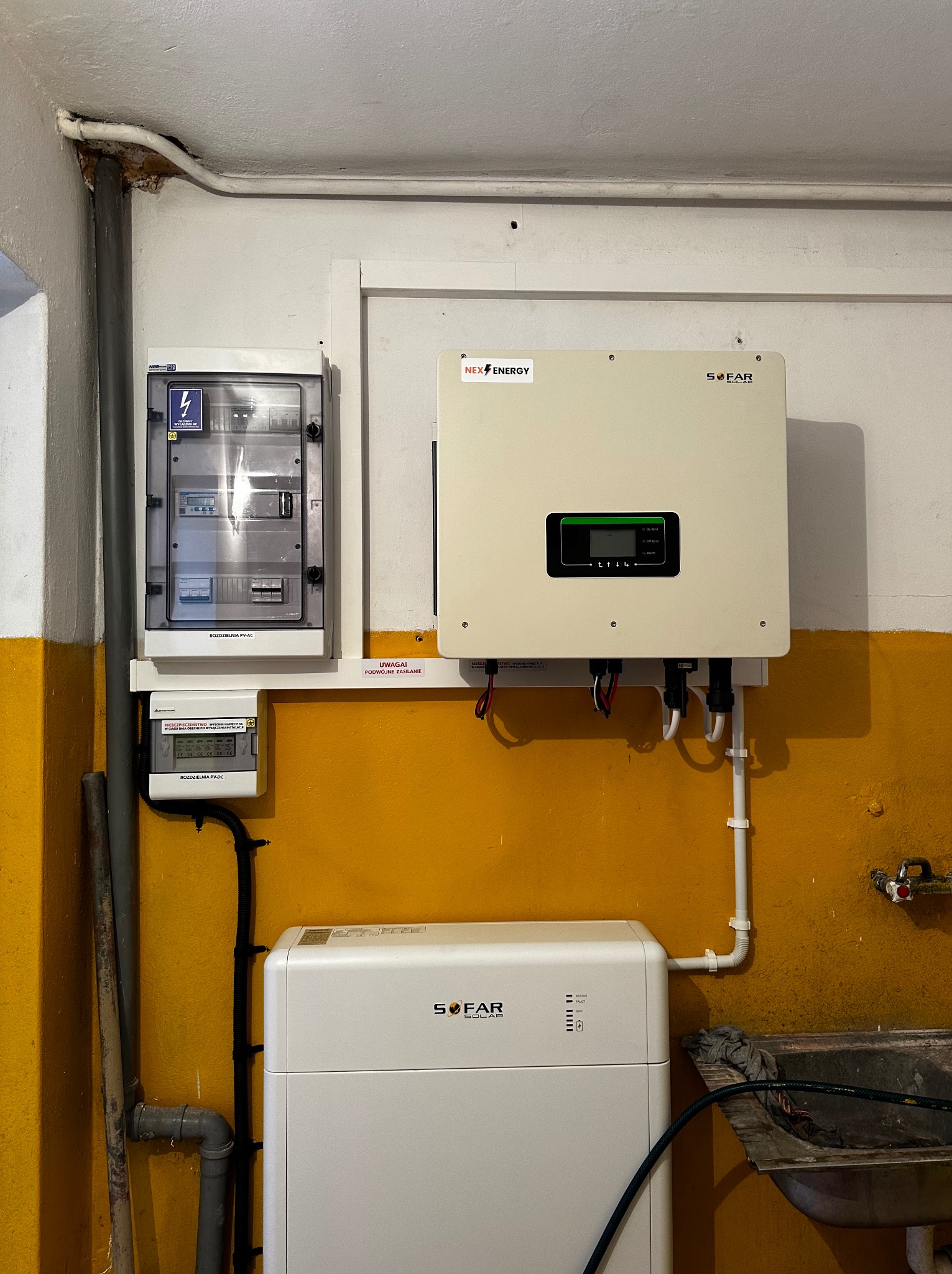 Instalacja z białym falownikiem i magazynem energii SOFAR oraz rozdzielnicą PV-AC i PV-DC na żółto-białej ścianie w pomieszczeniu technicznym.