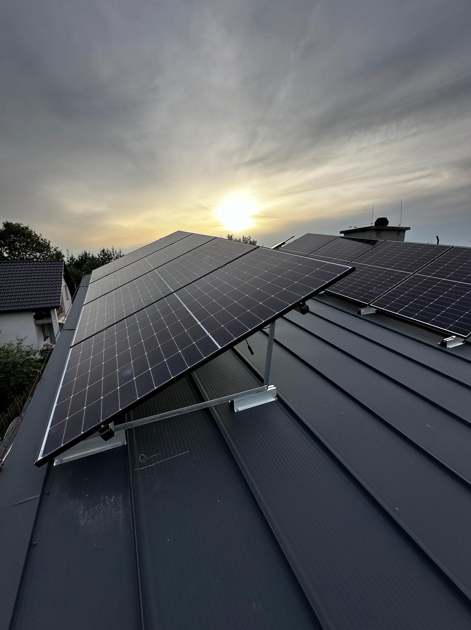 Panele słoneczne zamontowane na metalowym dachu z widocznym zachodzącym słońcem i pochmurnym niebem w tle.